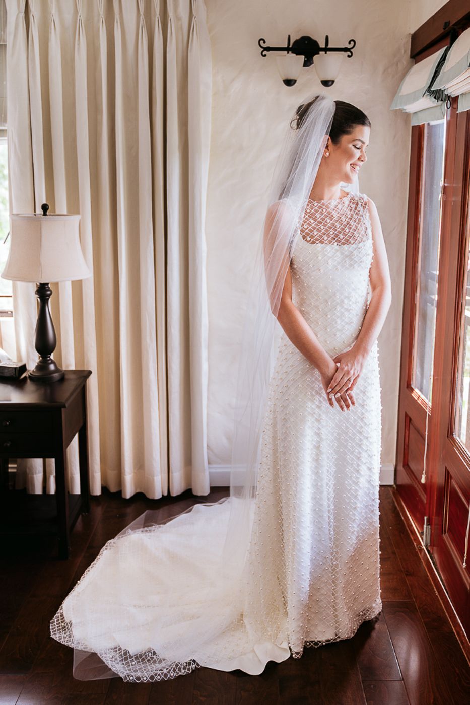 An elegant bride poses in a white lace wedding gown with a flowing train and veil in a sunlit room.