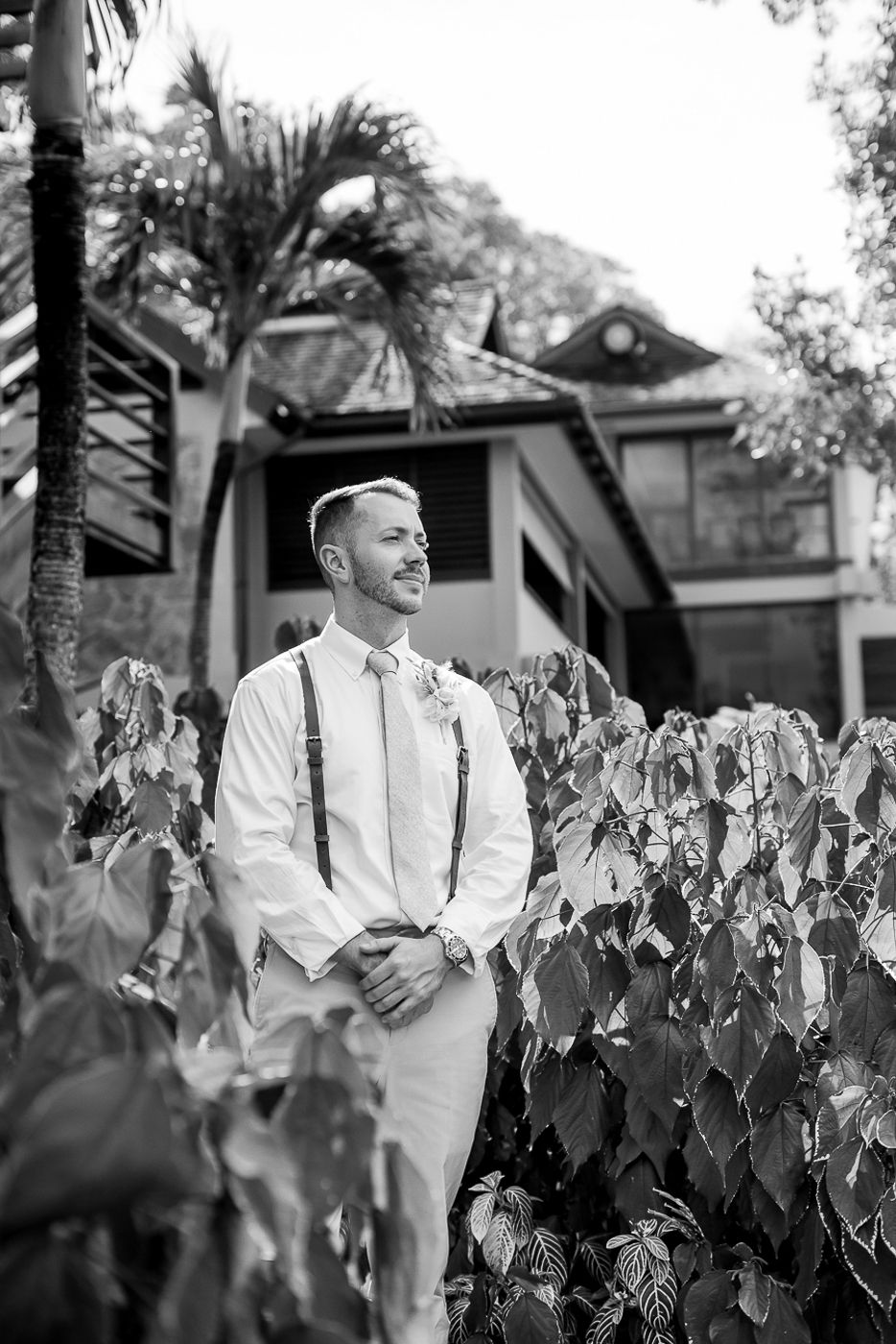 Black and white photo showing a groom in white attire and suspenders standing among tropical foliage.
