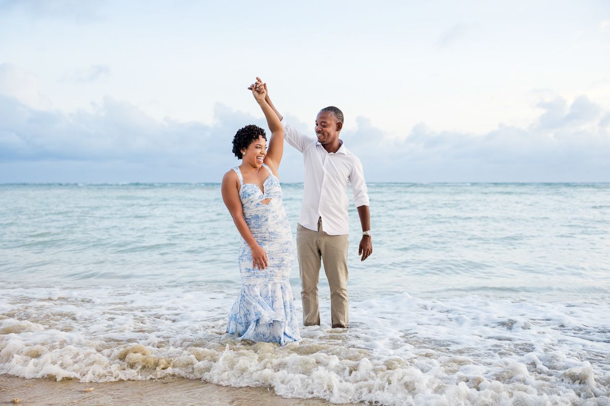 Happy couple dancing in the waves at the beach during golden hour with ocean and sky in background.