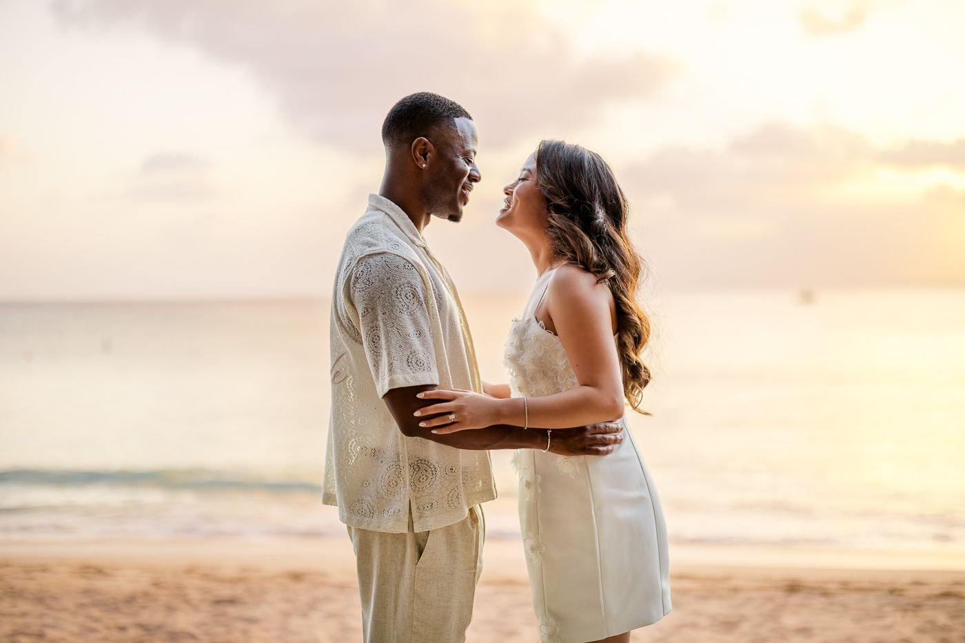 A couple shares an intimate moment on the beach during sunset wearing light colored outfits.