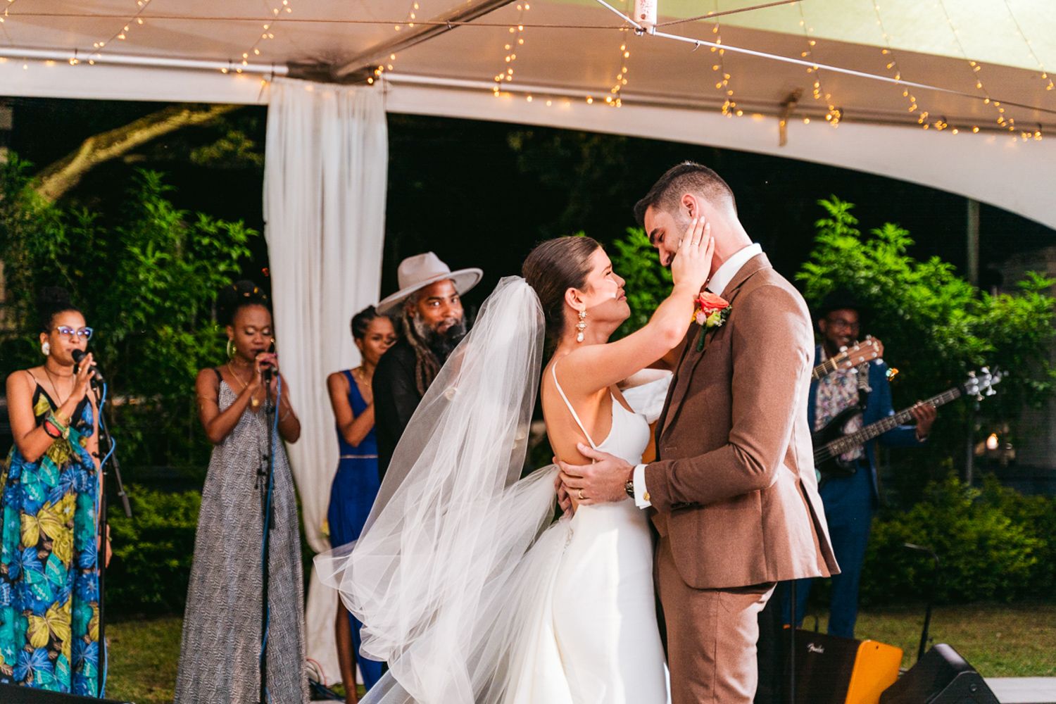 Wedding party dancing outdoors under string lights at evening reception in garden setting.
