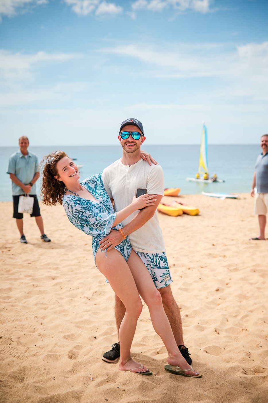 Playful couple dancing together on a sunny beach with sailboats in the background.