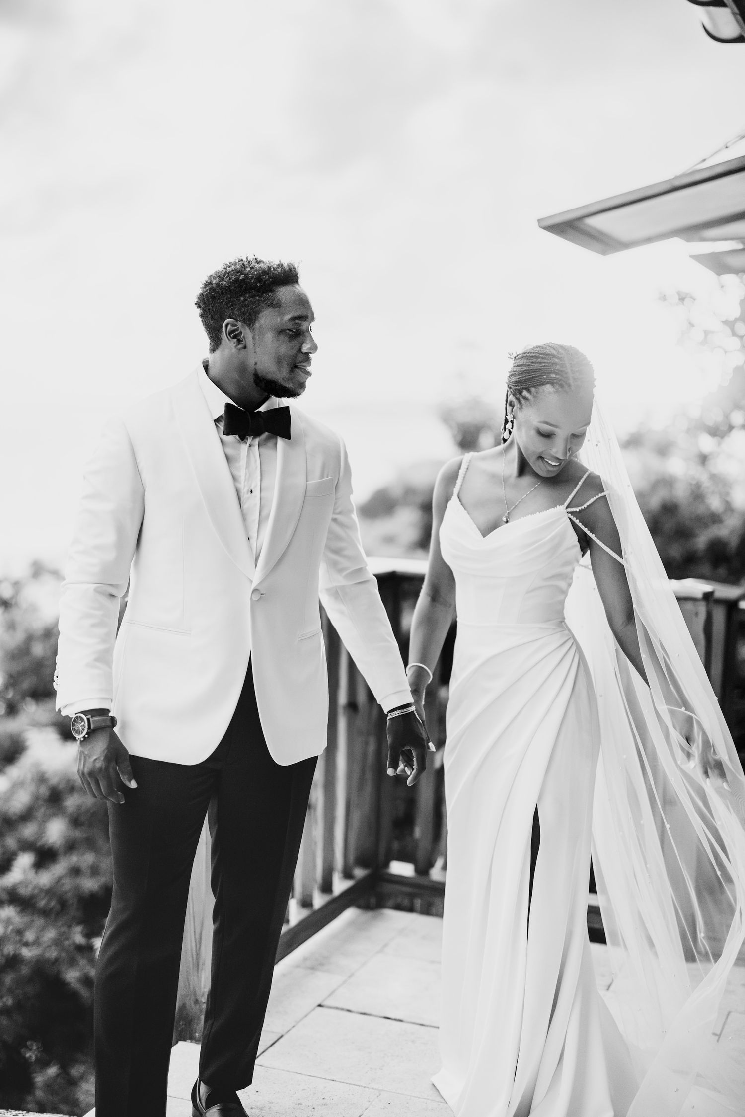 Black and white photo of a smiling bride and groom holding hands and walking together on a deck on their wedding day.