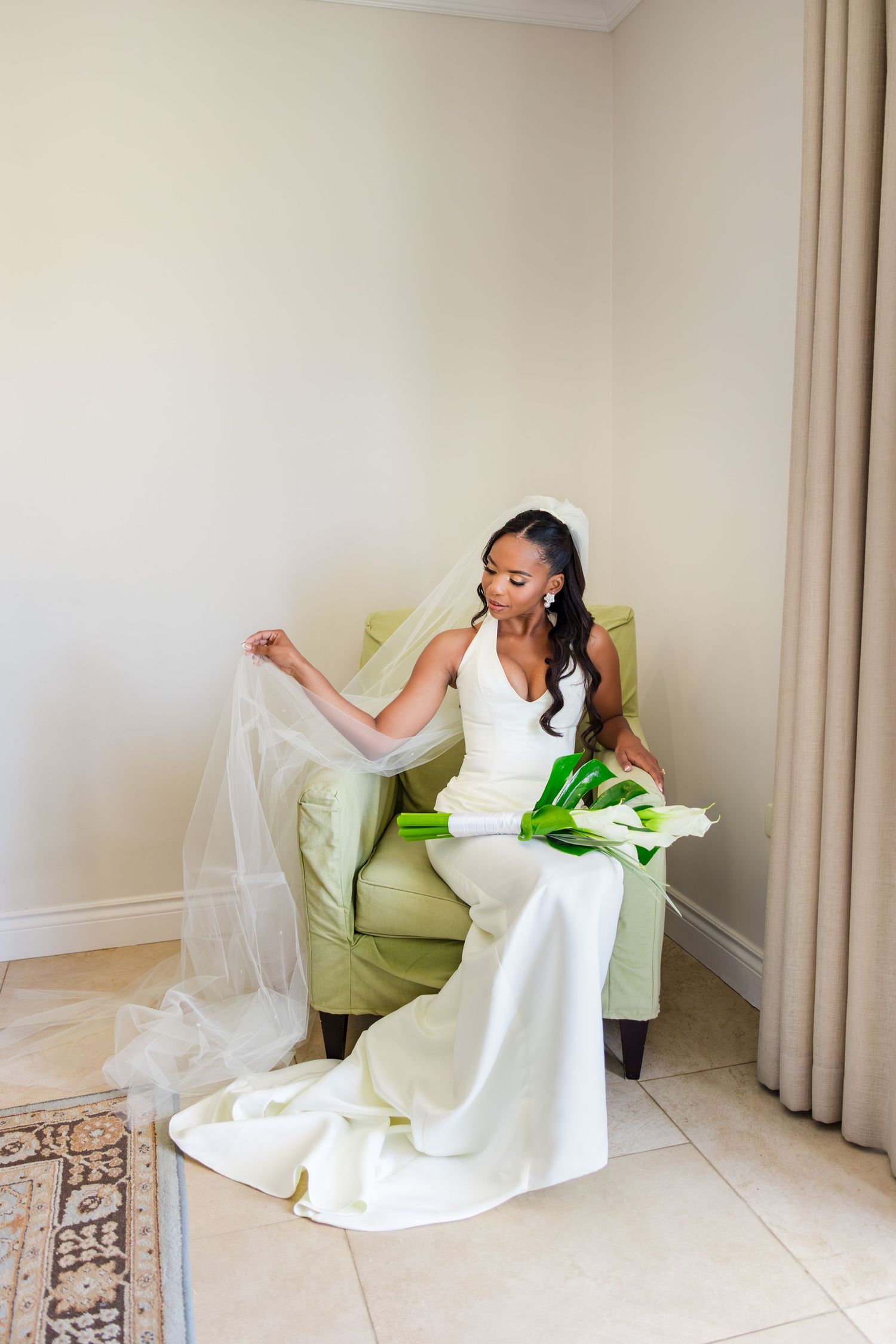 Elegant bride in white halter gown and veil sits in green chair holding calla lily bouquet in bright room.