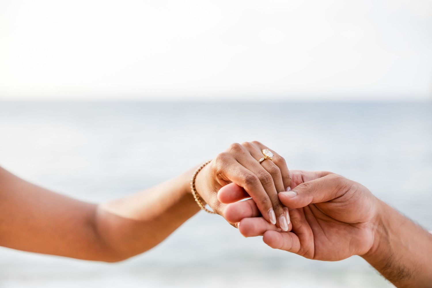 Couple holding hands tenderly on a beach, woman wearing a gold bracelet and engagement ring by the ocean.