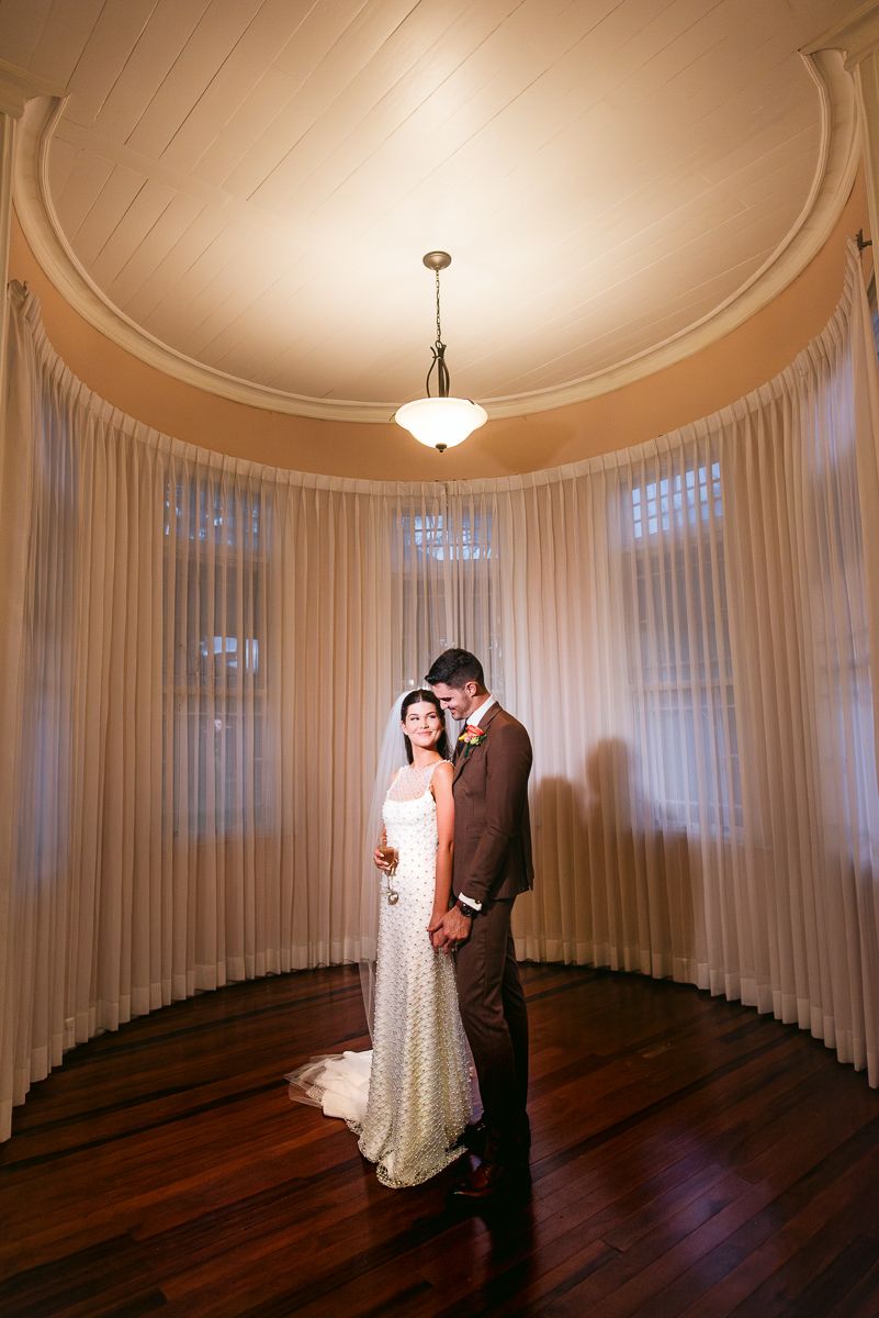 Elegant couple stands in circular room with white curtains, hardwood floors and domed ceiling with pendant light.