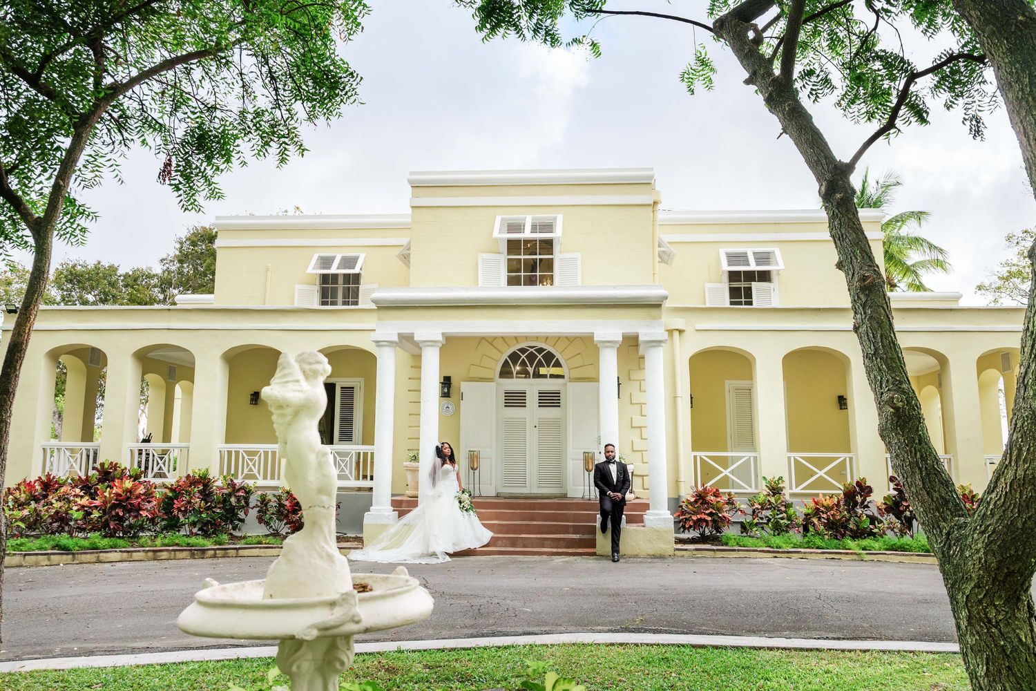 Bride and groom pose outside a grand yellow colonial mansion with white columns and lush tropical gardens.