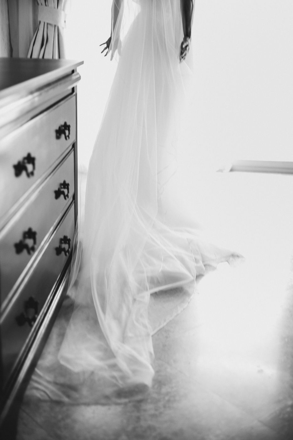 Black and white photo of a bride in a flowing white wedding gown standing near a dresser in soft natural light.
