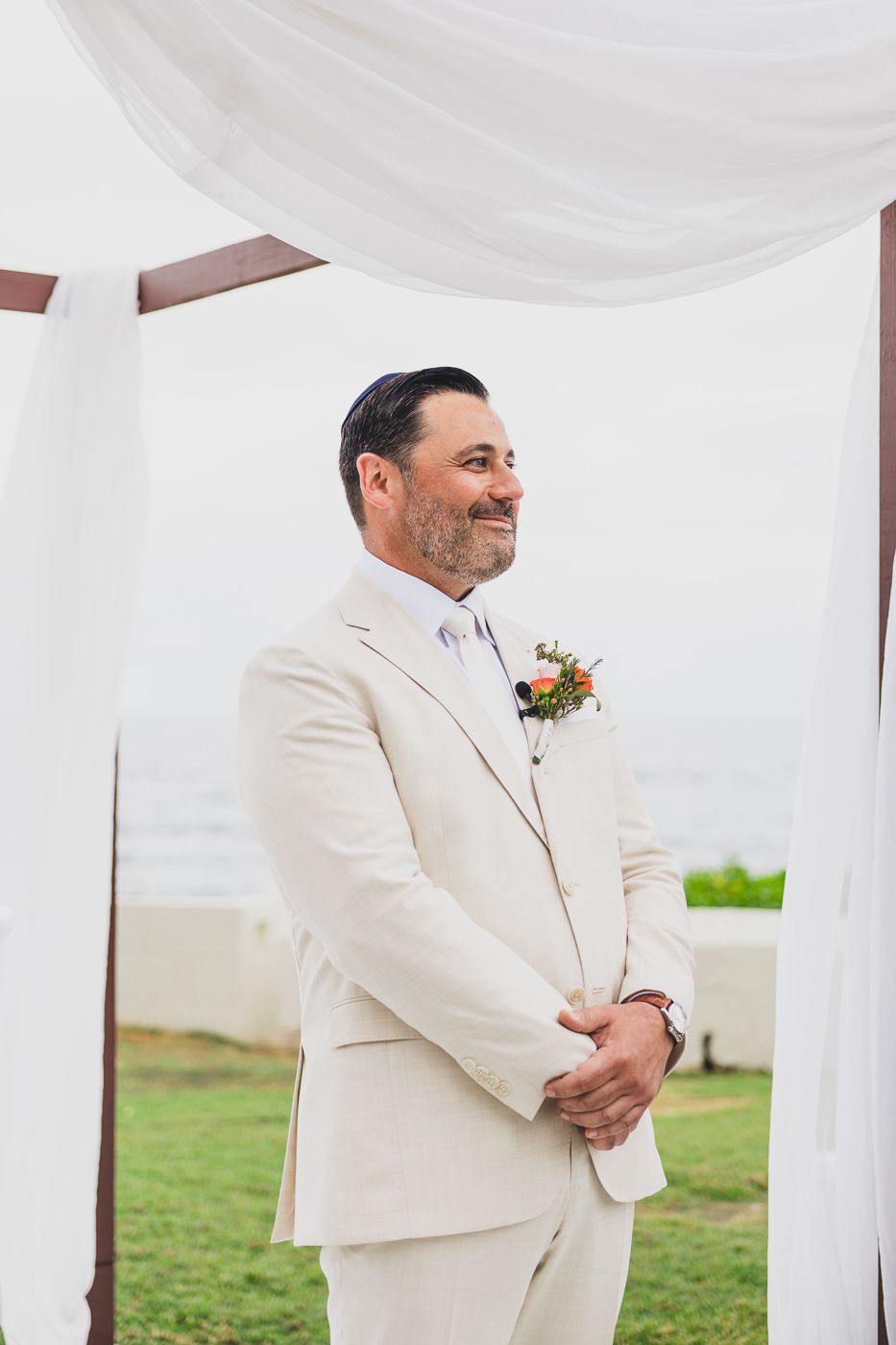 Smiling groom in cream suit with floral boutonniere stands under white draped arch at outdoor wedding ceremony.