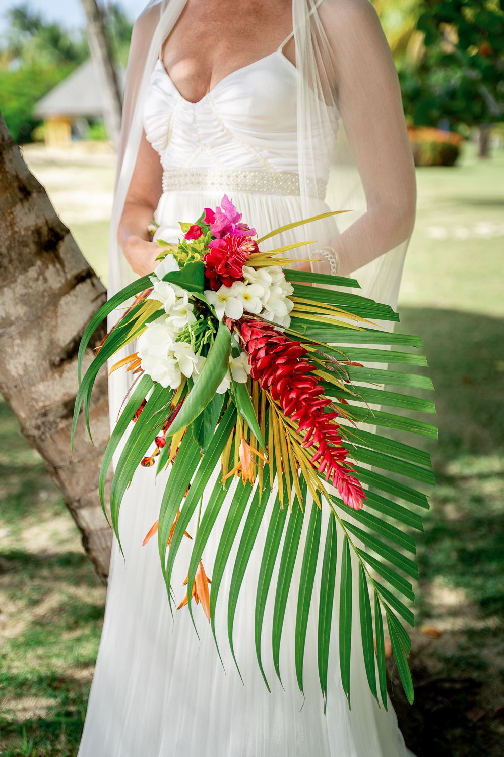 Bride in white gown holding tropical wedding bouquet with palm leaves, red ginger, and white orchids outdoors.
