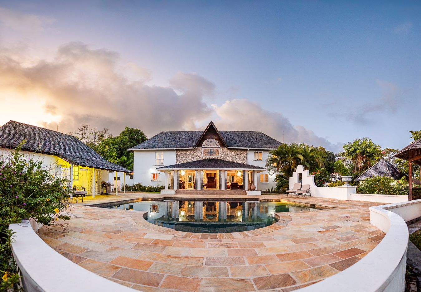 Luxury Caribbean villa with illuminated interiors, reflecting pool, and stone patio at sunset.