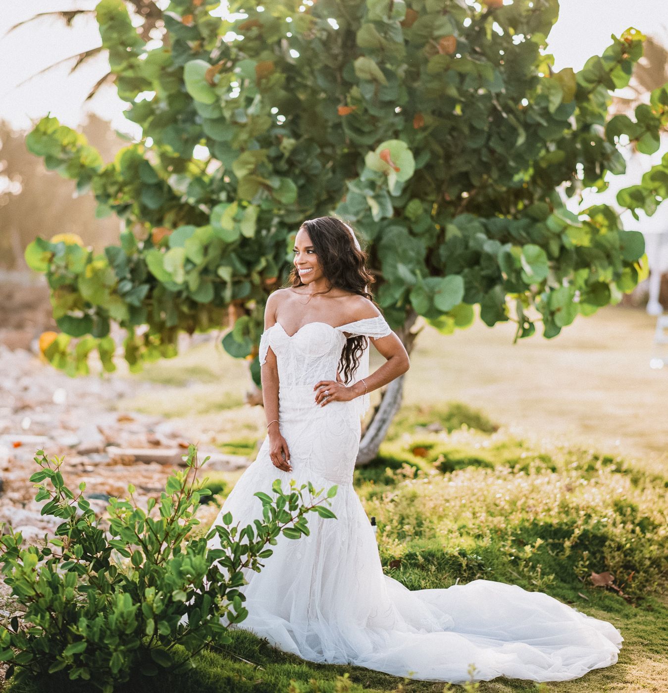 A bride in a fitted mermaid wedding gown poses gracefully in a sunlit garden setting.