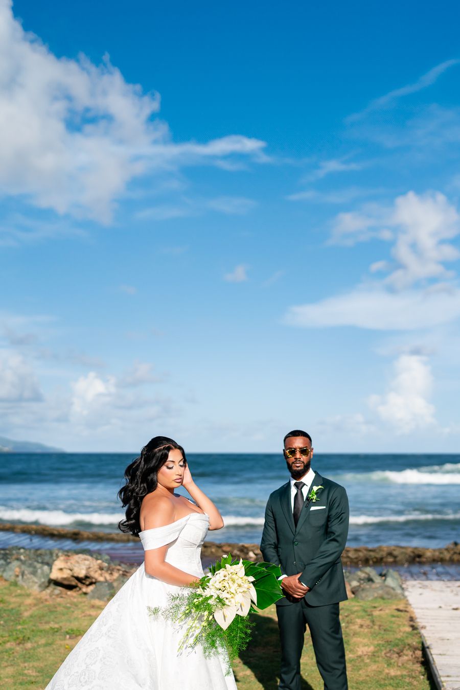 Wedding couple poses along rocky oceanfront with waves and blue sky in the background.