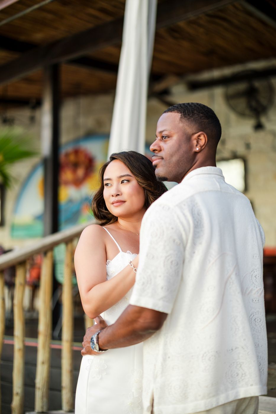 A couple in matching white outfits embrace while standing on a rustic porch with decorative columns.