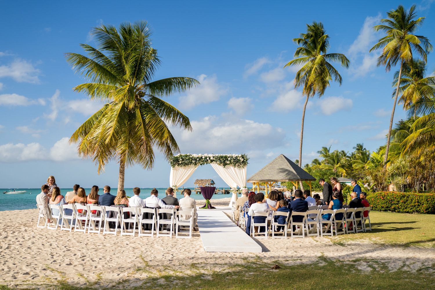 Beach wedding ceremony setup with white chairs and palm trees along tropical shoreline.