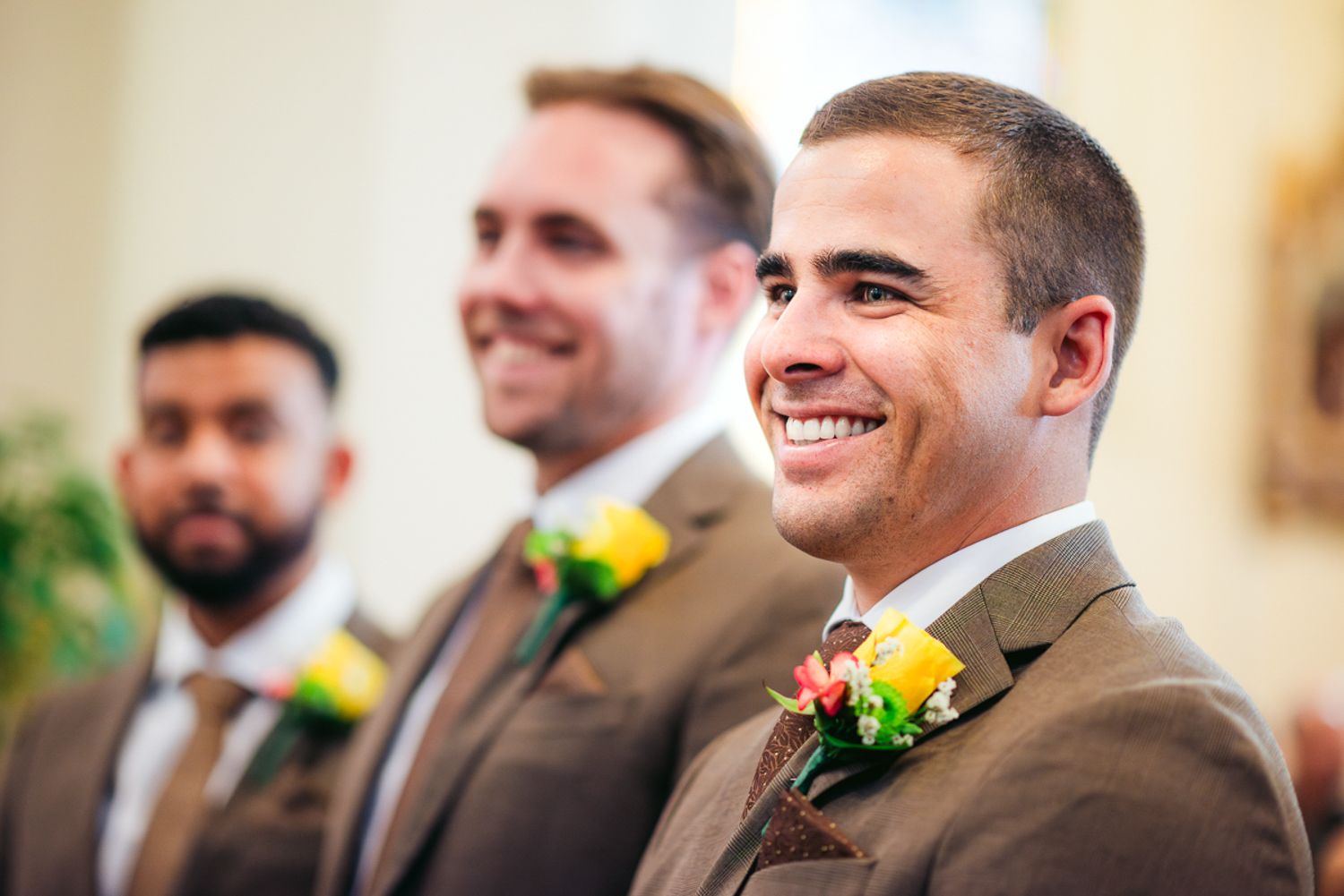Groomsmen in brown suits smile during wedding ceremony wearing yellow rose boutonnieres.
