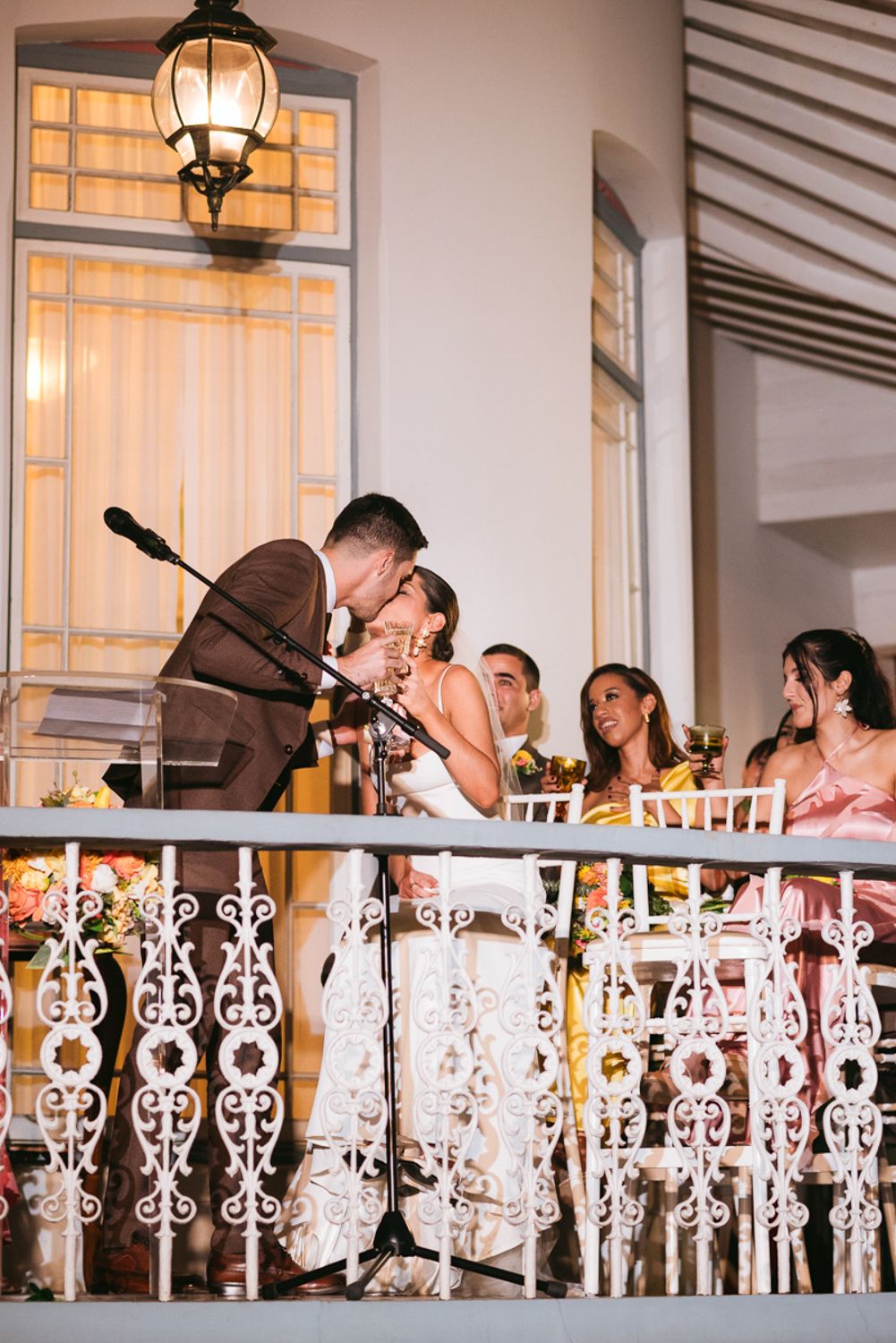A group gathers on an elegant balcony with ornate white railings at a formal event as lanterns glow overhead.