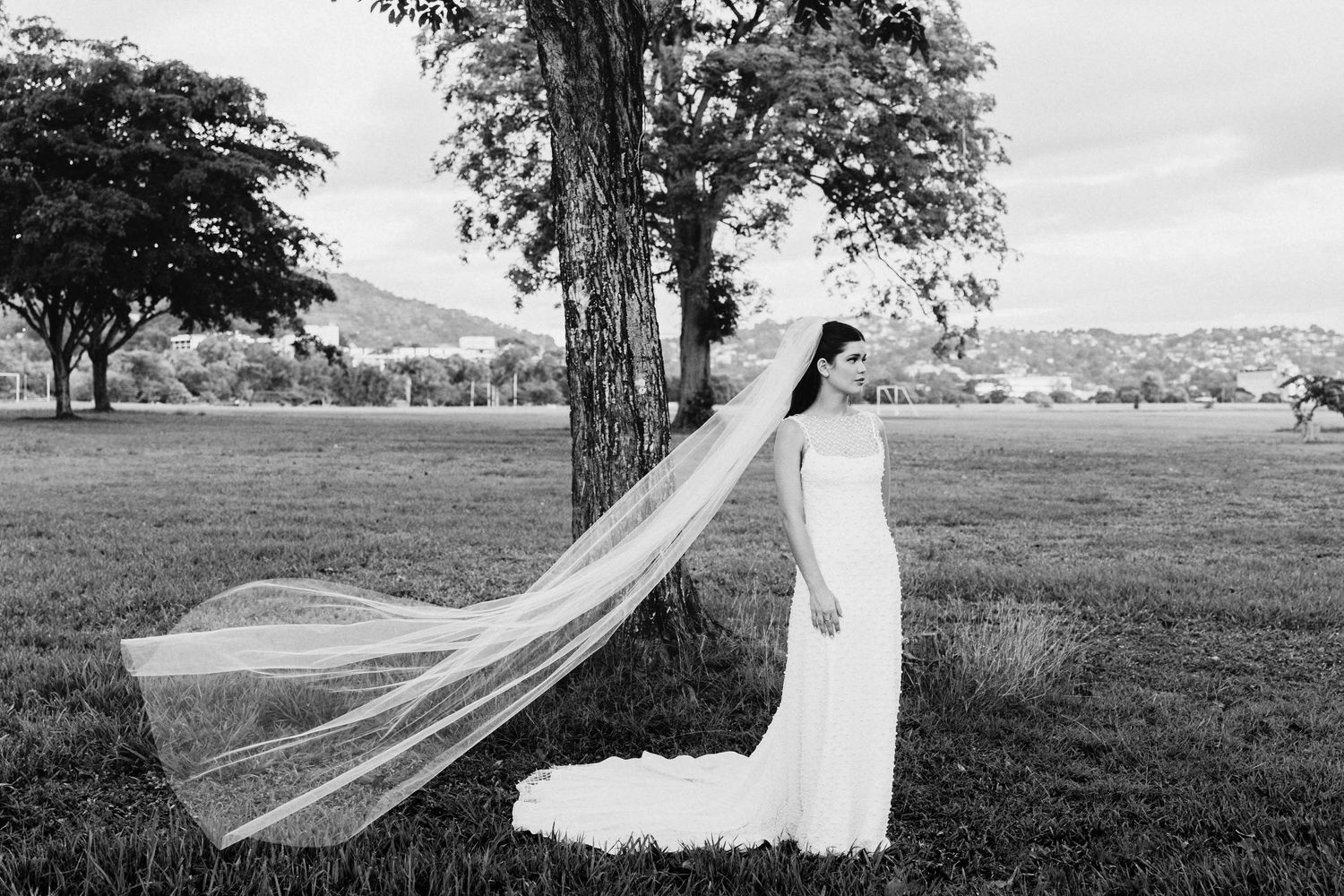 A flowing wedding veil catches the wind in a dramatic black and white photograph on a grassy hillside.