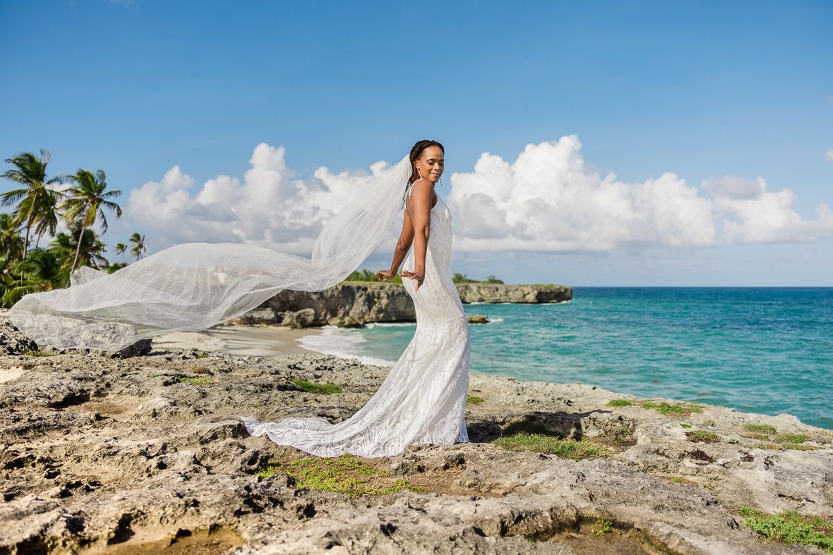Bride in white wedding dress with flowing veil on rocky tropical coastline with turquoise ocean and palm trees.