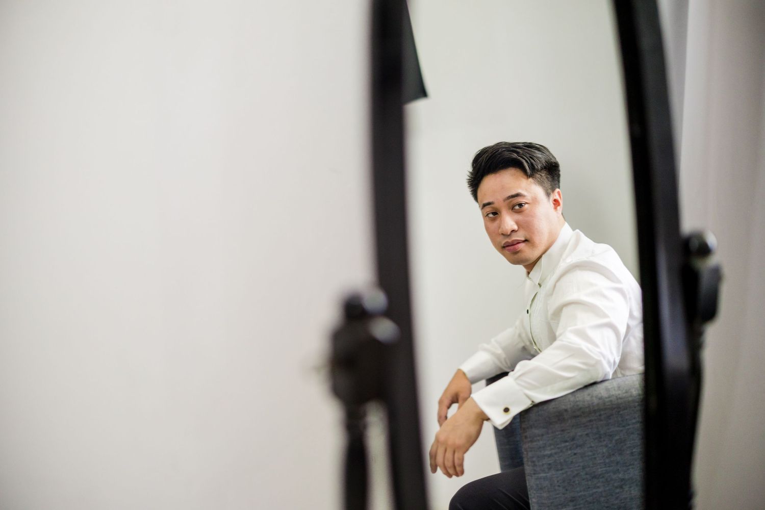 Young Asian man in white shirt sitting confidently in a studio photography setting with professional lighting equipment.