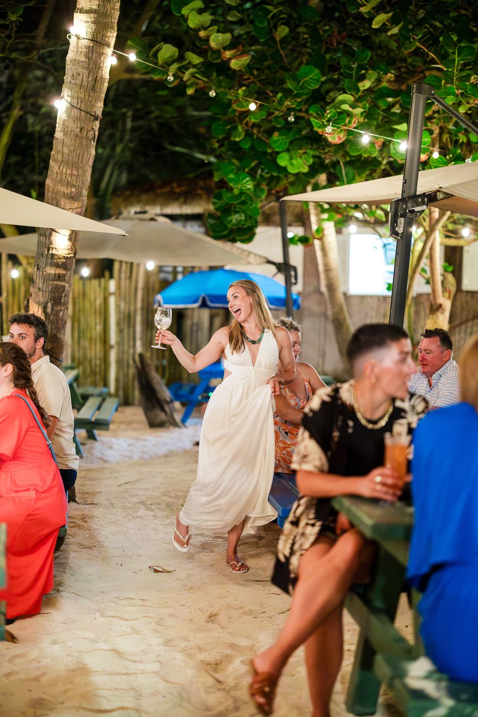 Woman in white dress dancing joyfully at an outdoor beach party at night, holding a wine glass under string lights.