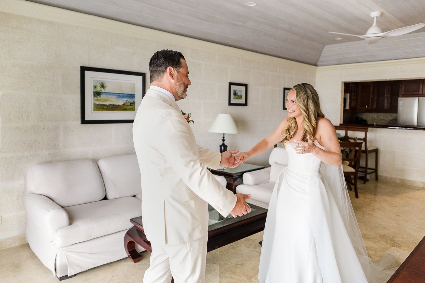 Bride and groom sharing a first look moment in a bright hotel suite, both dressed in elegant white wedding attire.