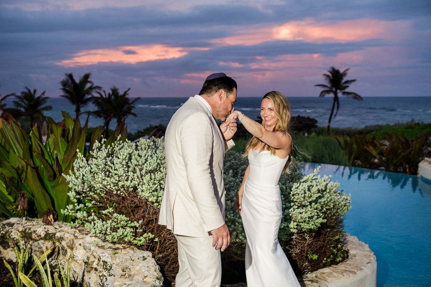 Bride and groom share a romantic sunset moment by a pool with ocean views and palm trees at a tropical wedding.