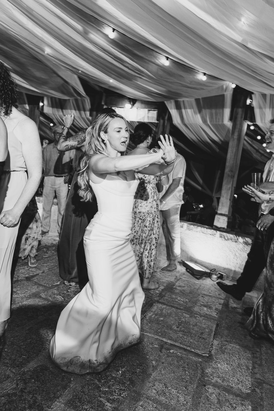 Bride in white gown dancing joyfully with guests under draped ceiling lights at wedding reception.