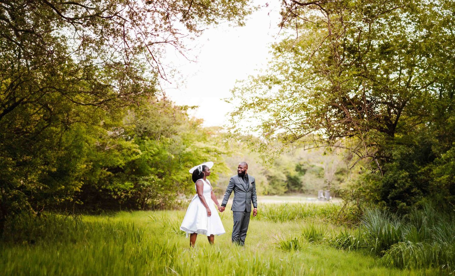 A couple in wedding attire holds hands in a lush green meadow surrounded by tall trees on their special day.