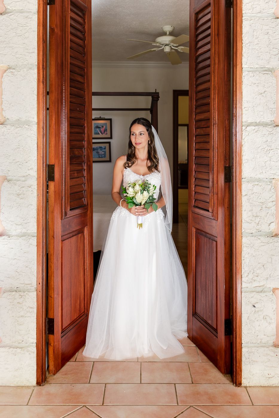 Elegant bride in a white tulle gown holding a bouquet standing in wooden doorway of stone building.