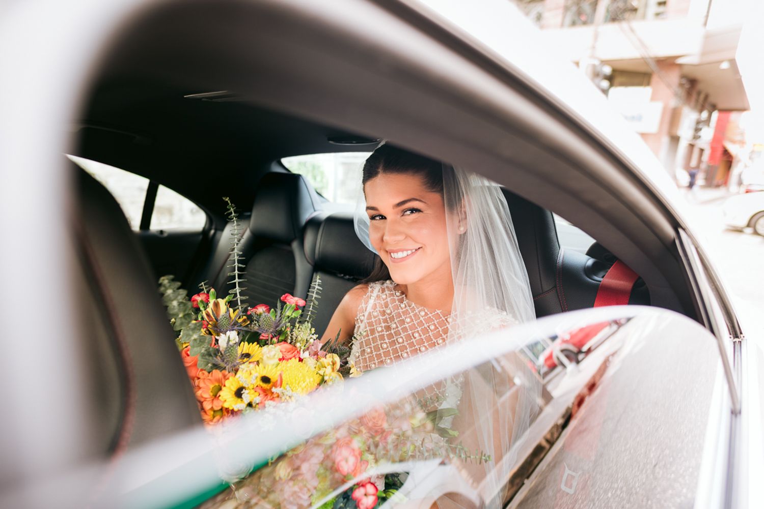 A bride sits inside a car wearing a white wedding veil while holding flowers.