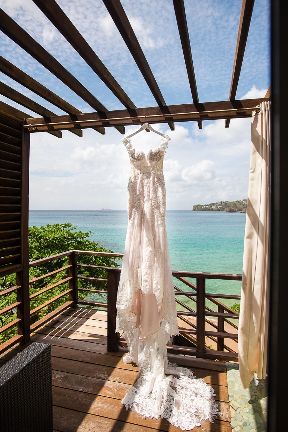 White lace wedding dress hanging on a tropical balcony overlooking turquoise ocean waters.