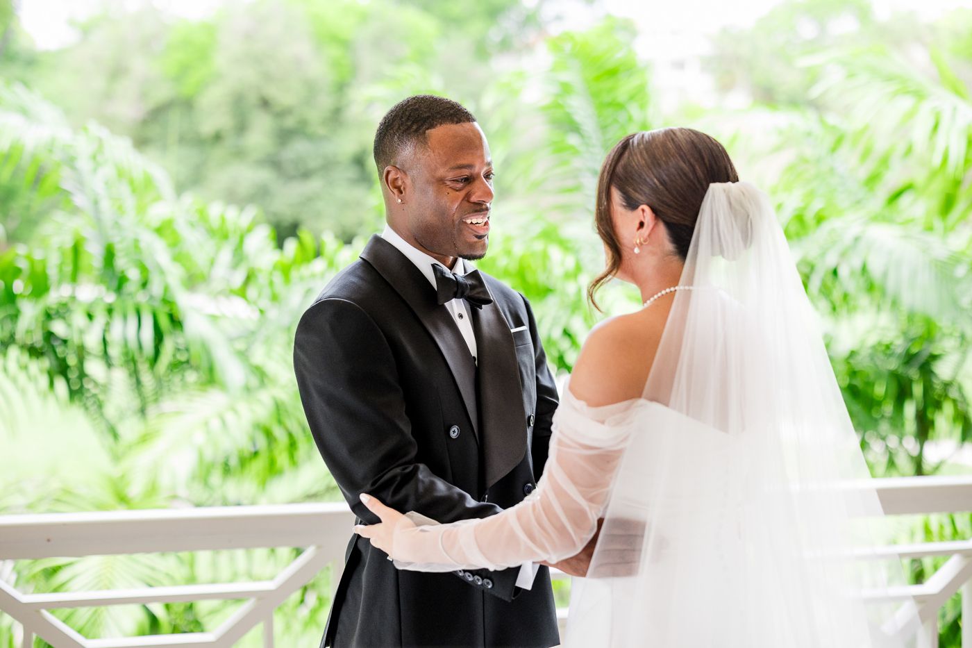 An intimate first look moment unfolds on a bright porch with tropical landscaping in the background.