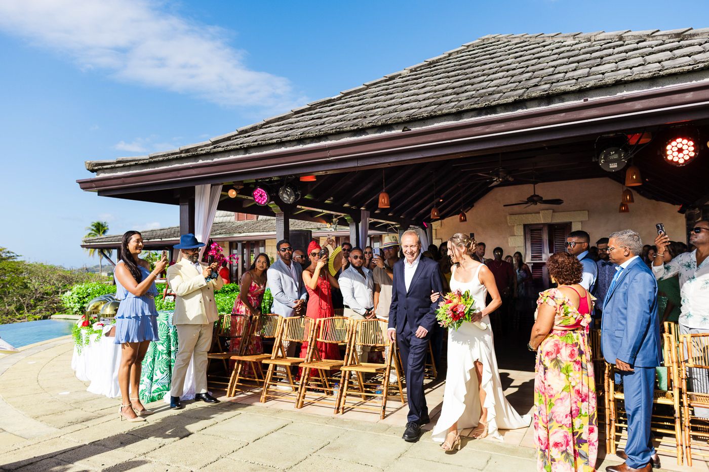 Wedding guests gather on covered patio overlooking tropical ocean view.