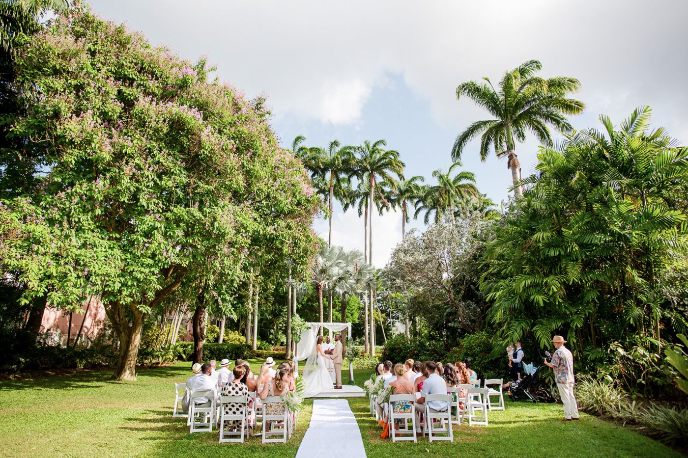 Outdoor wedding venue setup with white chairs and aisle runner lined by towering palm trees.