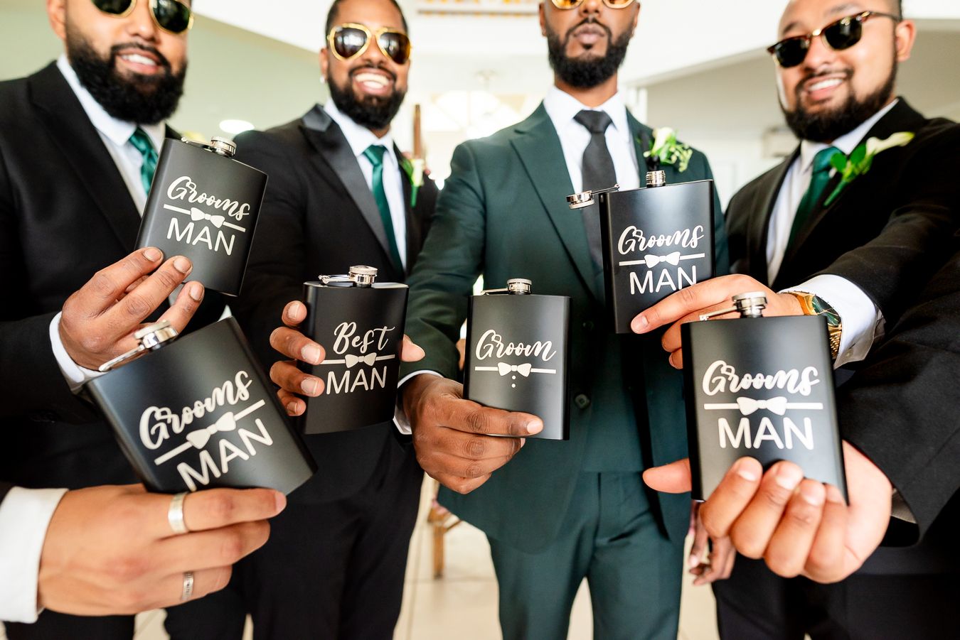Groomsmen in sunglasses and suits holding matching black flasks for a group photo.