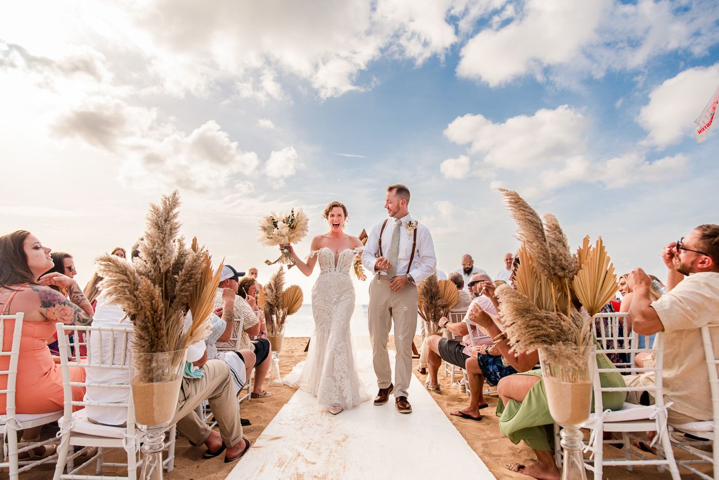 Wedding ceremony guests seated in white chairs outdoors under a blue sky with fluffy clouds.