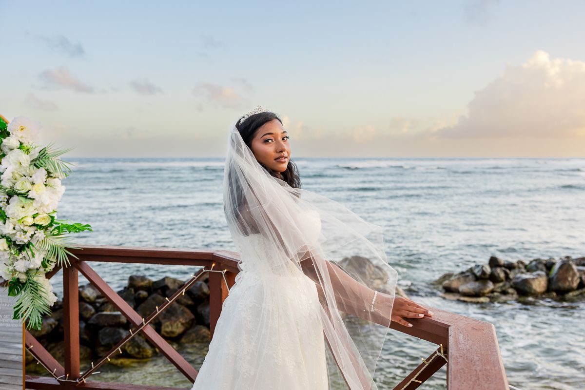 A bride in a flowing white wedding dress poses gracefully against a scenic ocean backdrop at sunset.