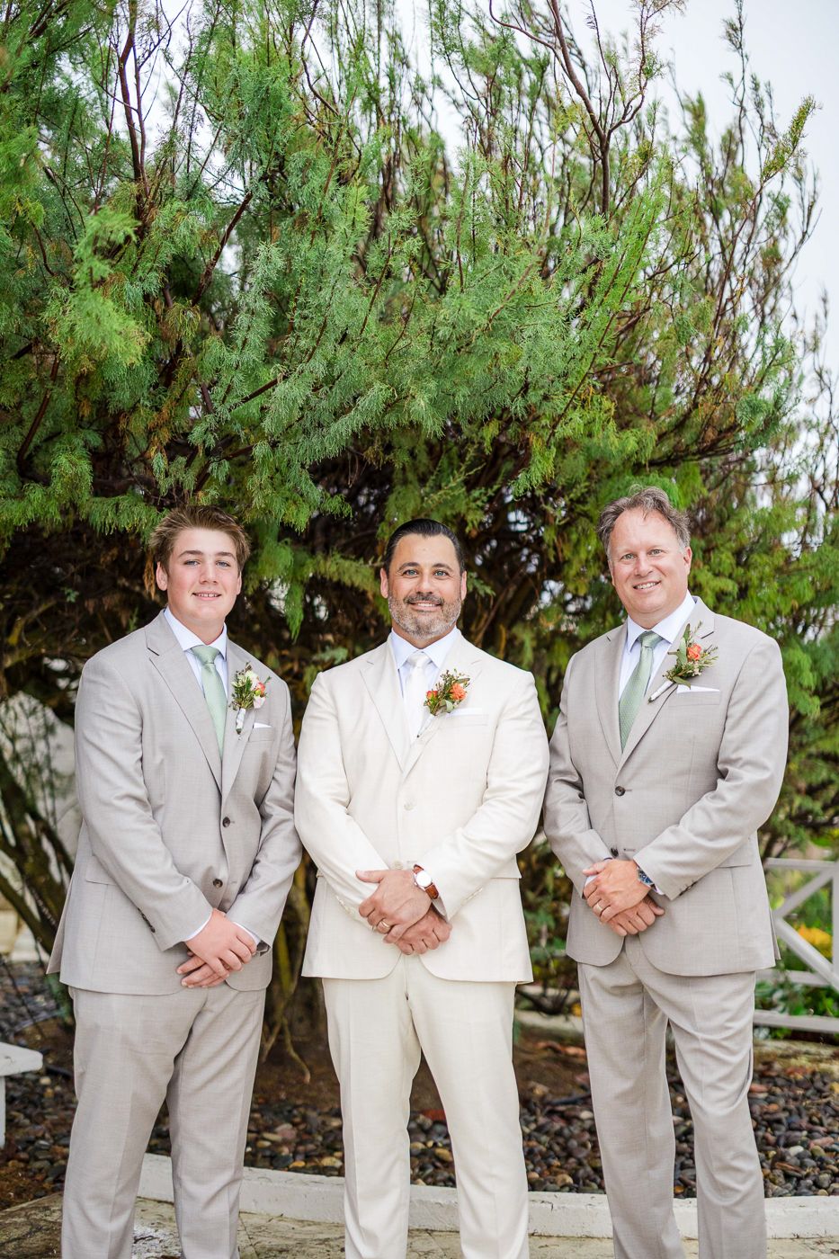 Three men in suits pose outdoors at a wedding, with the groom in white flanked by two groomsmen in tan suits.