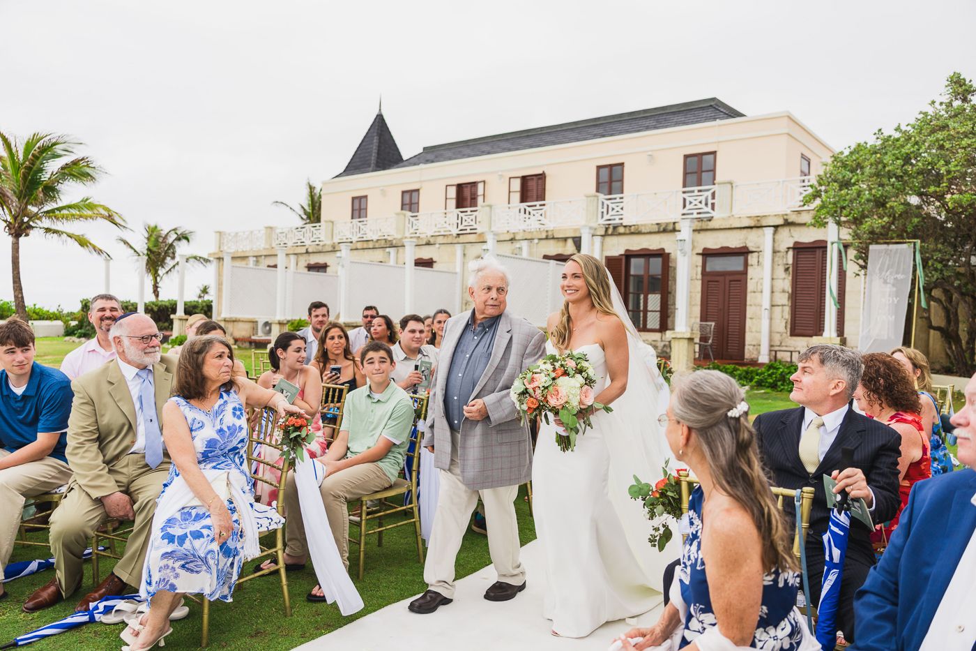 Bride walks down the aisle with escort at outdoor wedding ceremony near tropical venue with guests seated.