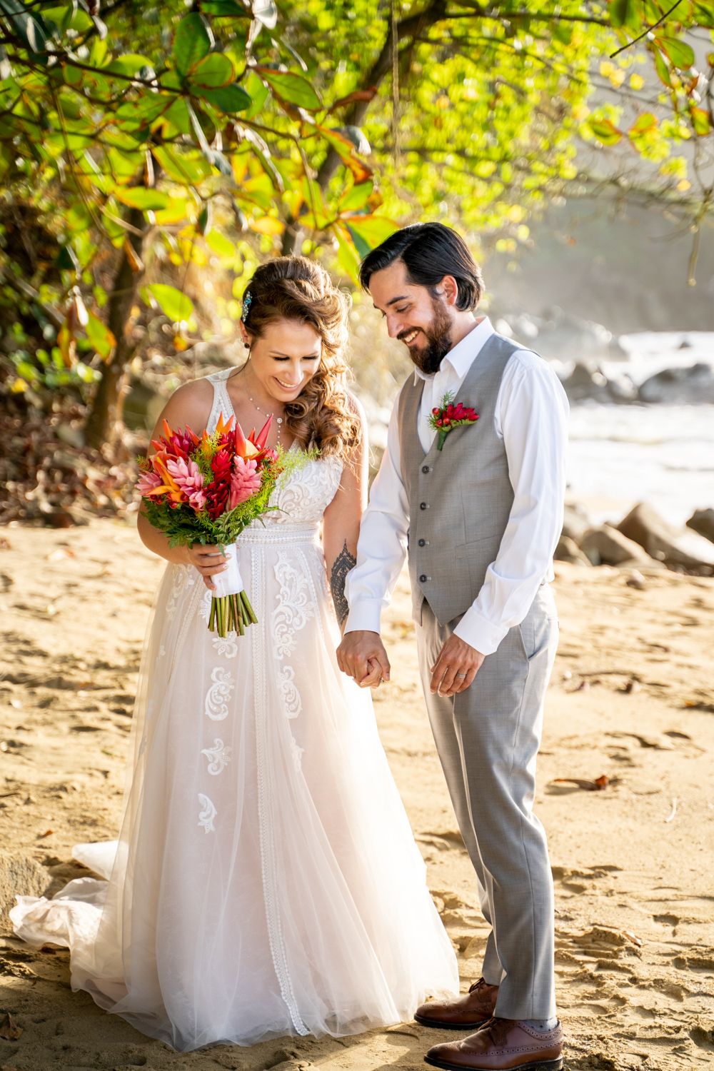 Bride and groom hold hands on a sandy beach, she holds a tropical bouquet, both smiling in wedding attire.