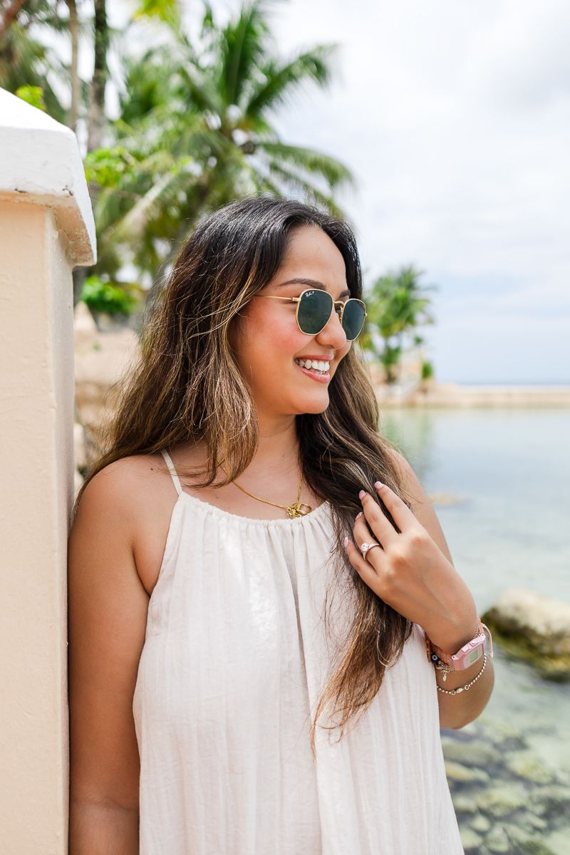 A person in a white summer dress and sunglasses smiles while standing near a tropical beach with palm trees.