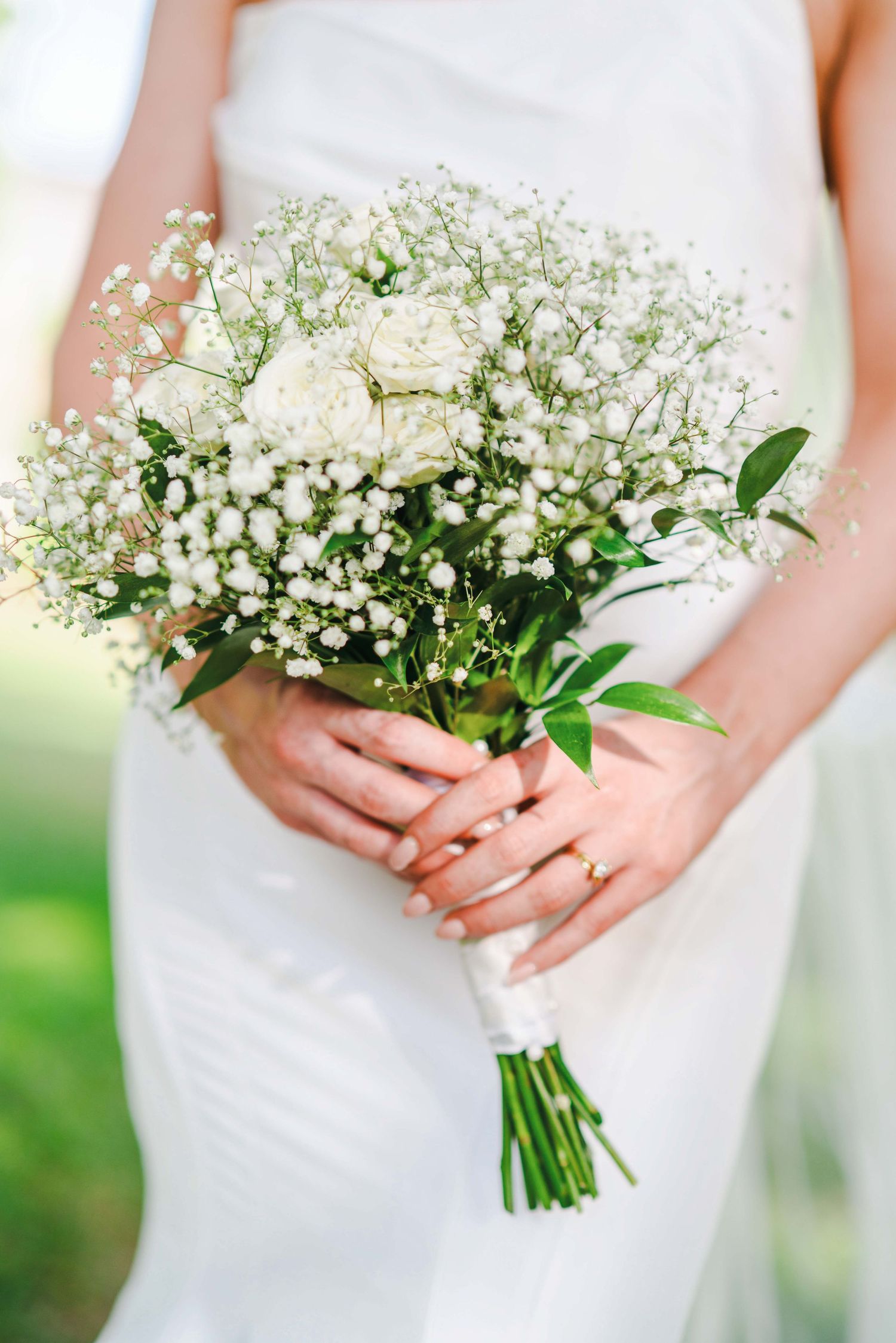 Delicate white rose and baby's breath bridal bouquet held by hands wearing wedding ring.