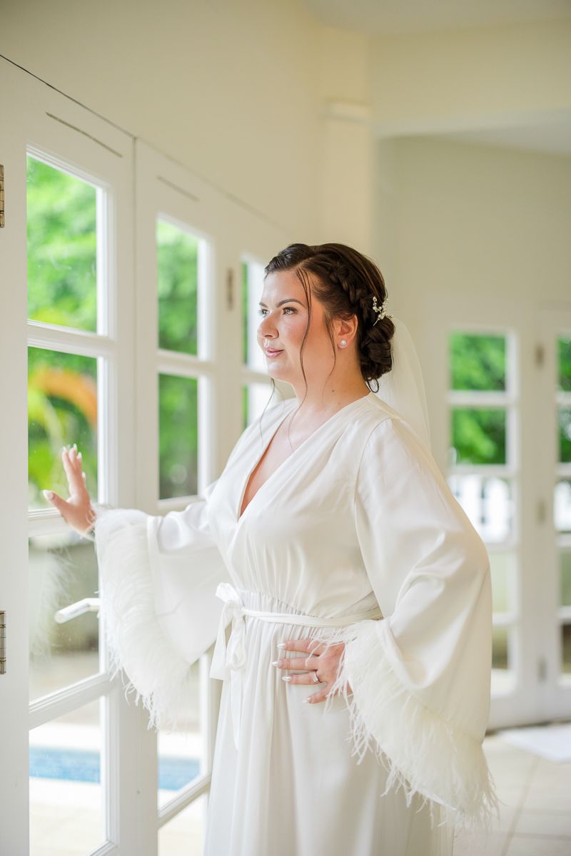 Bride in white feathered robe gazing out French doors, hair styled with accessories, in bright elegant room.