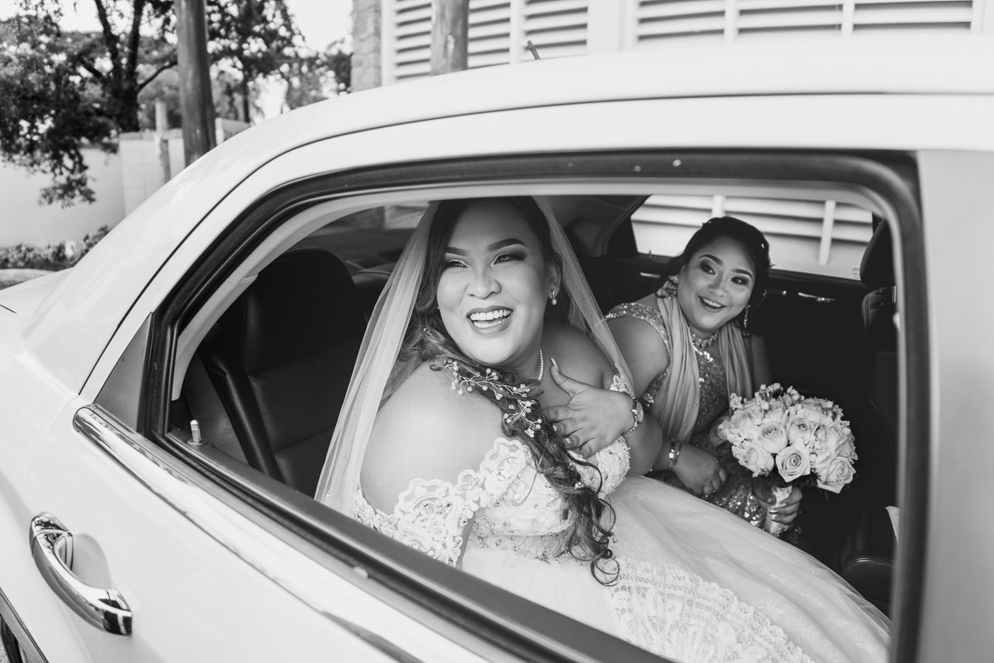 Bride in wedding dress smiling from car window with bridesmaid holding bouquet in black and white photo.