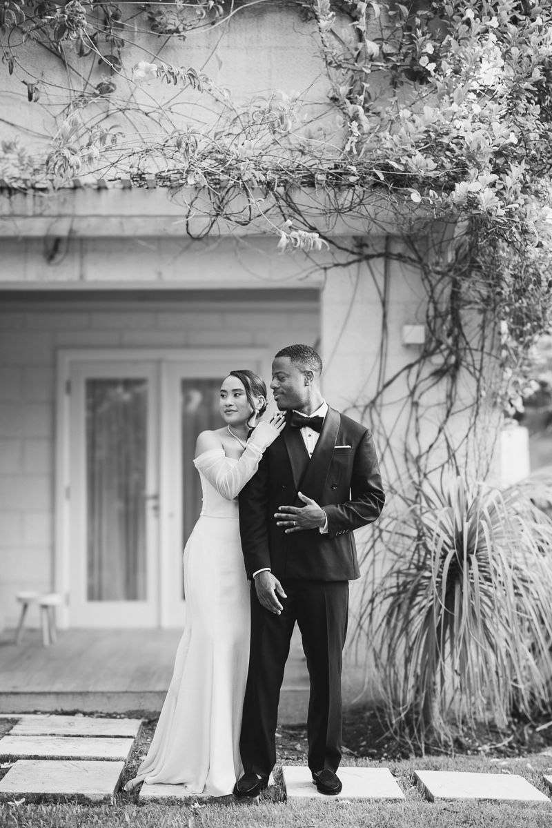 Black and white photo of a bride and groom posing outdoors in front of a vine-covered building on their wedding day.