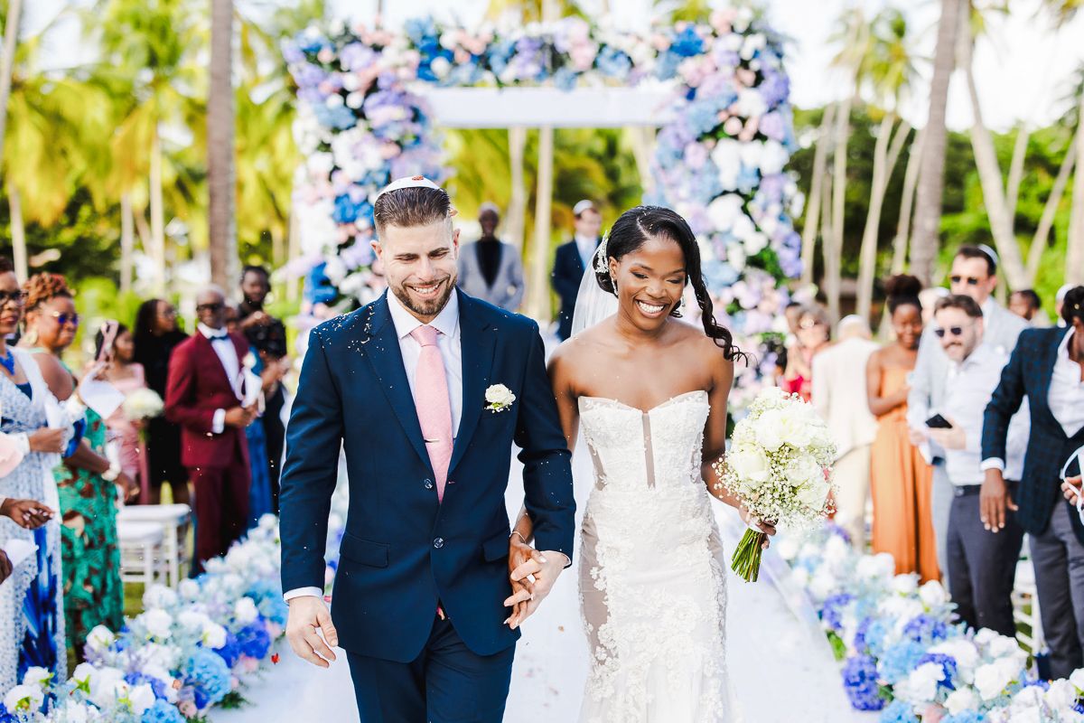 Bride and groom walk down aisle at outdoor tropical wedding with blue floral arch and guests celebrating.