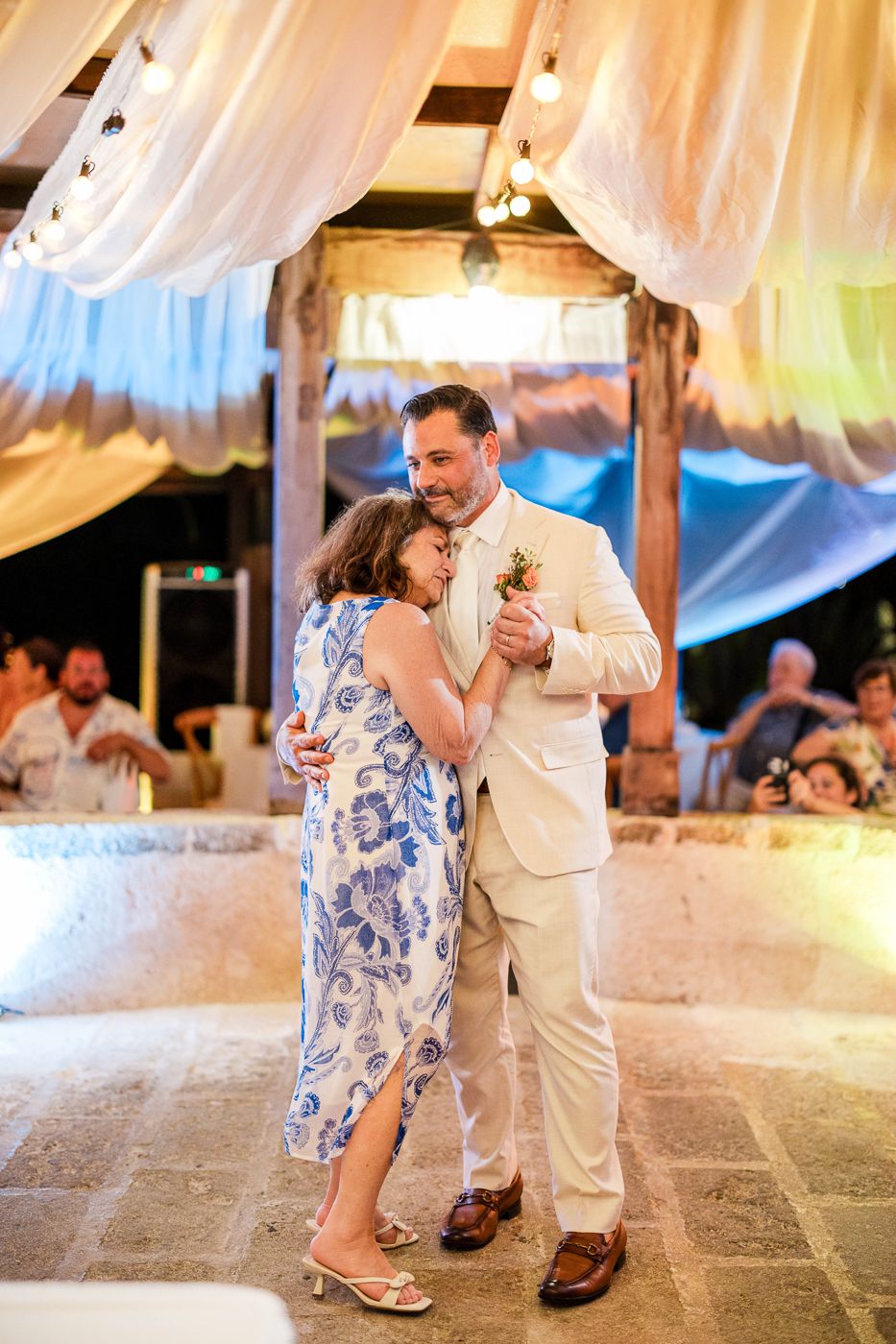 Groom in ivory suit shares mother-son dance with woman in blue floral dress at elegant outdoor wedding reception.