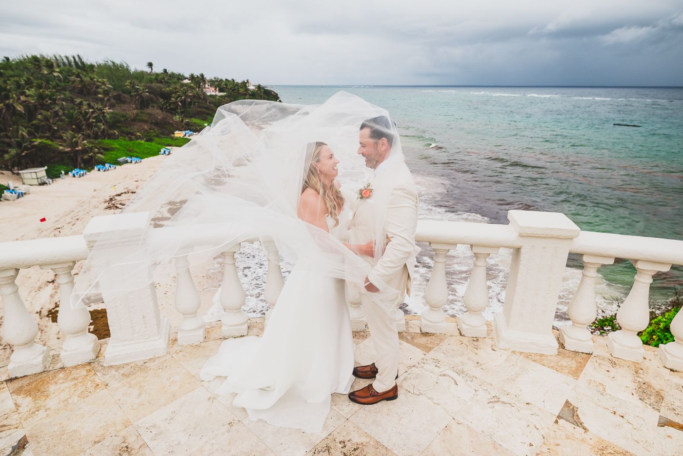 Bride and groom embrace on white stone terrace overlooking turquoise Caribbean sea, wedding veil flowing in the wind.
