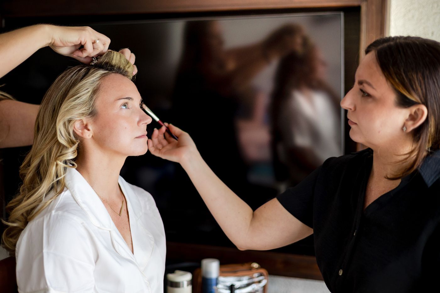 Makeup artist applying cosmetics to a blonde bride in a white robe during wedding preparation.
